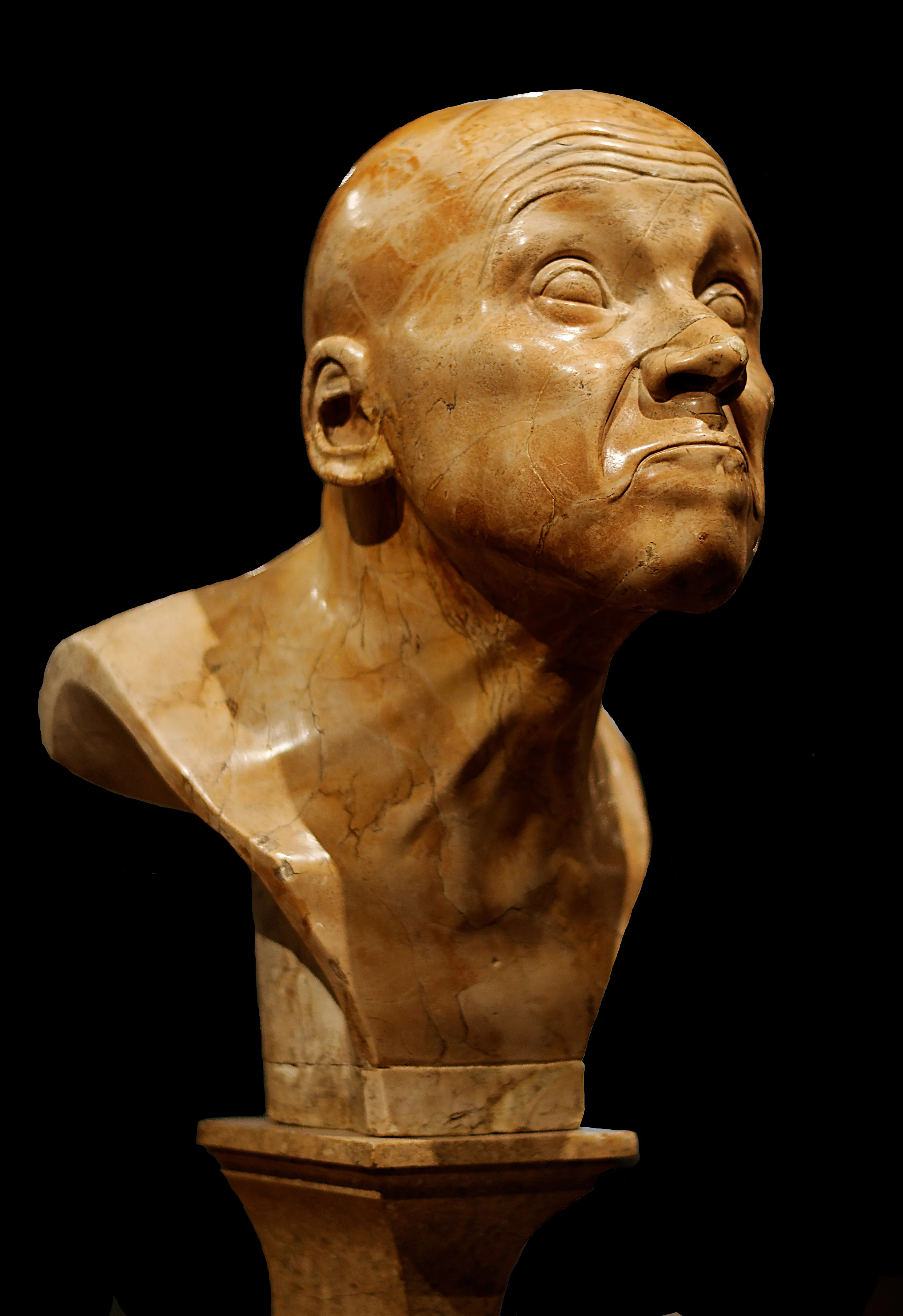 Een marmeren buste van een man met een overdreven, grimasende uitdrukking, tentoongesteld op een sokkel tegen een zwarte achtergrond.