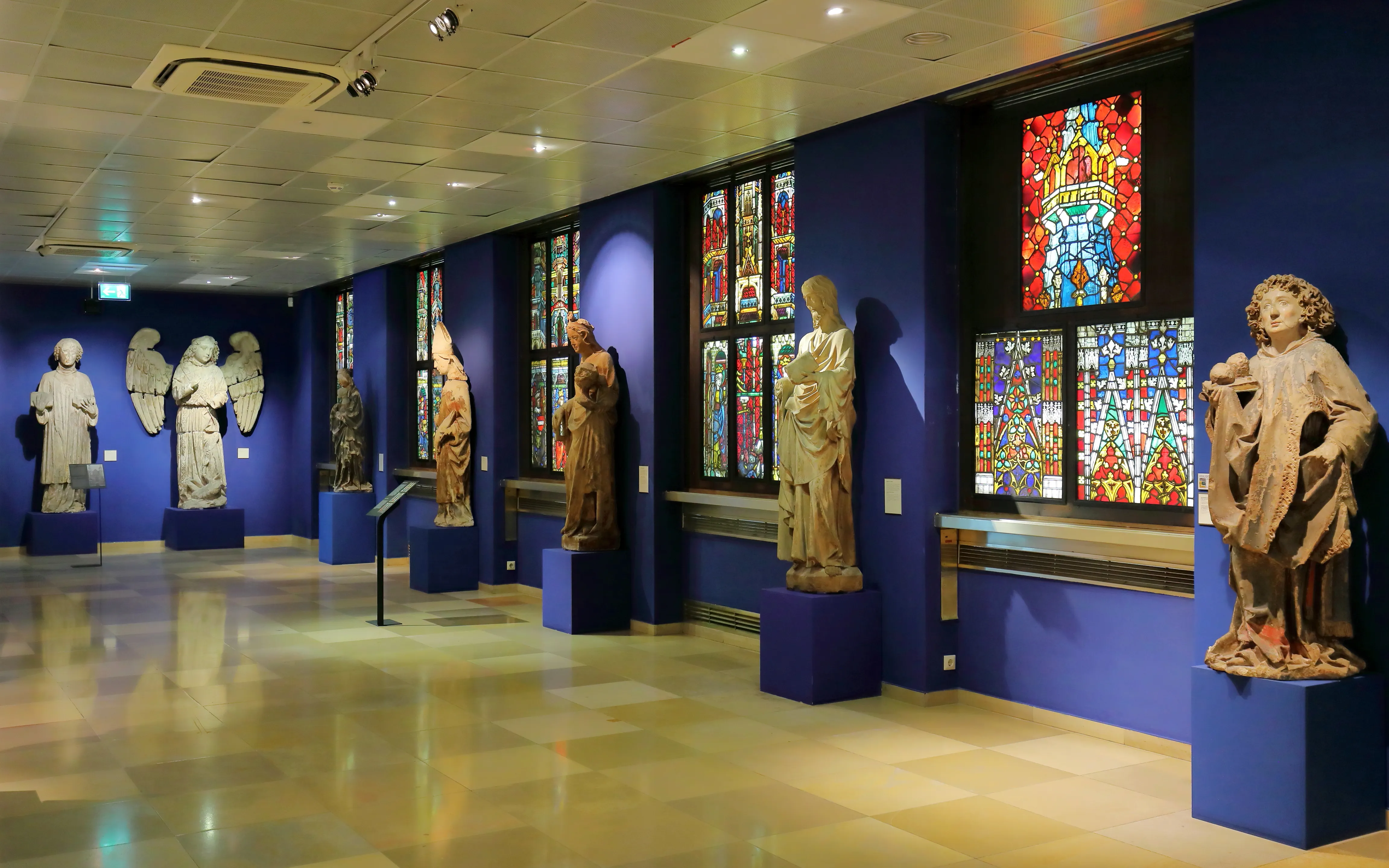 Interieur van het Wien Museum met beelden op blauwe sokkels en glas-in-loodramen tegen blauwe muren