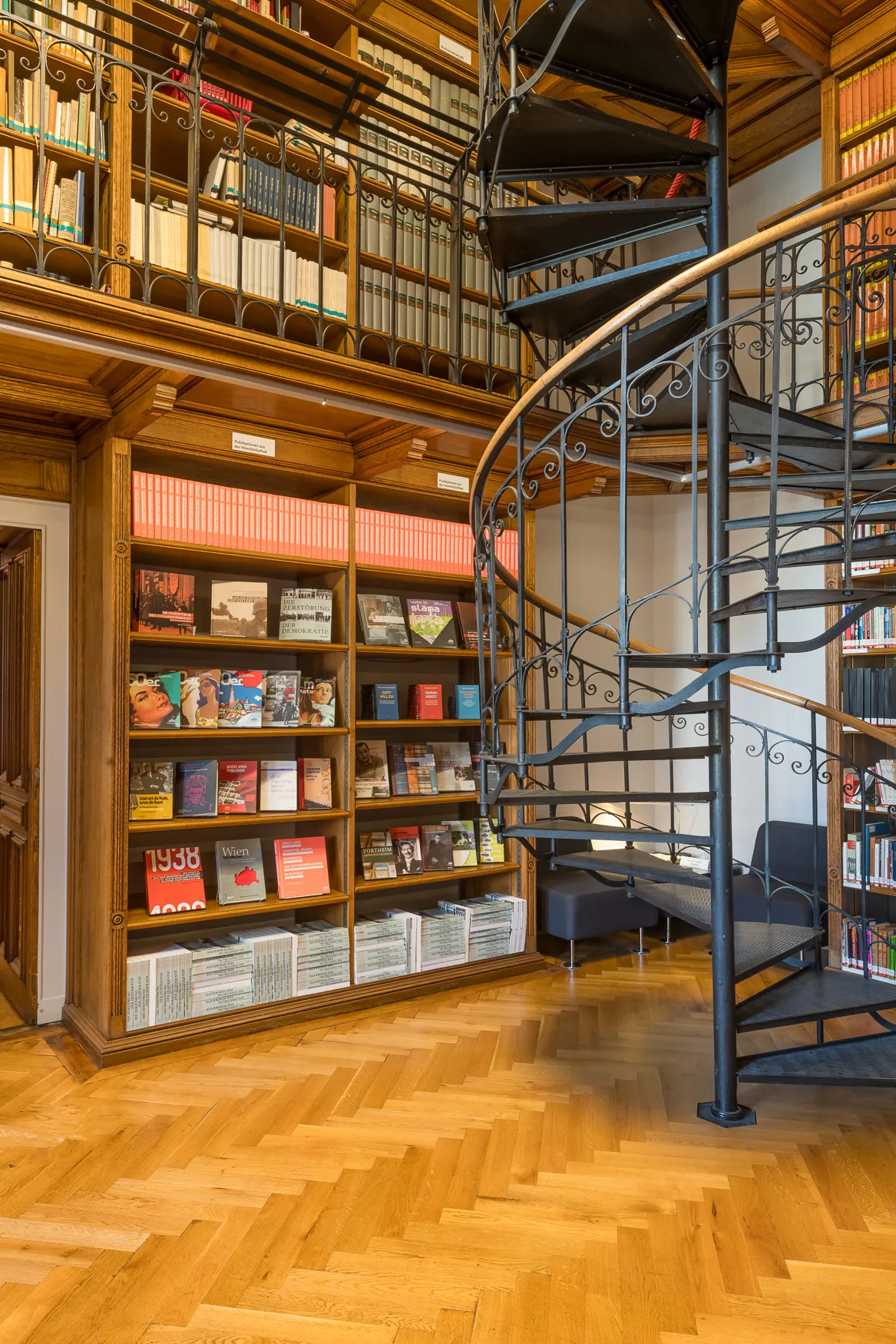 Wnętrze biblioteki z czarnymi metalowymi schodami kręconymi, drewnianymi półkami na książki i podłogą z drewna ułożoną w jodełkę