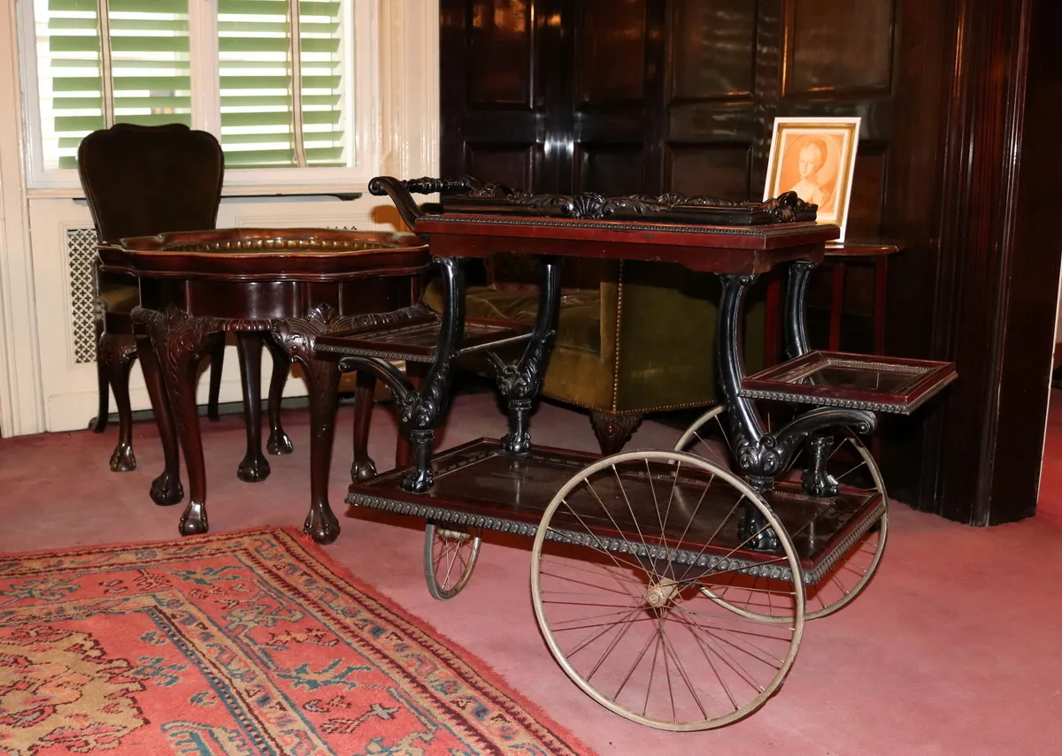 A vintage wooden serving cart with metal wheels and ornate legs, displayed in a room with dark wood paneling and a patterned rug.