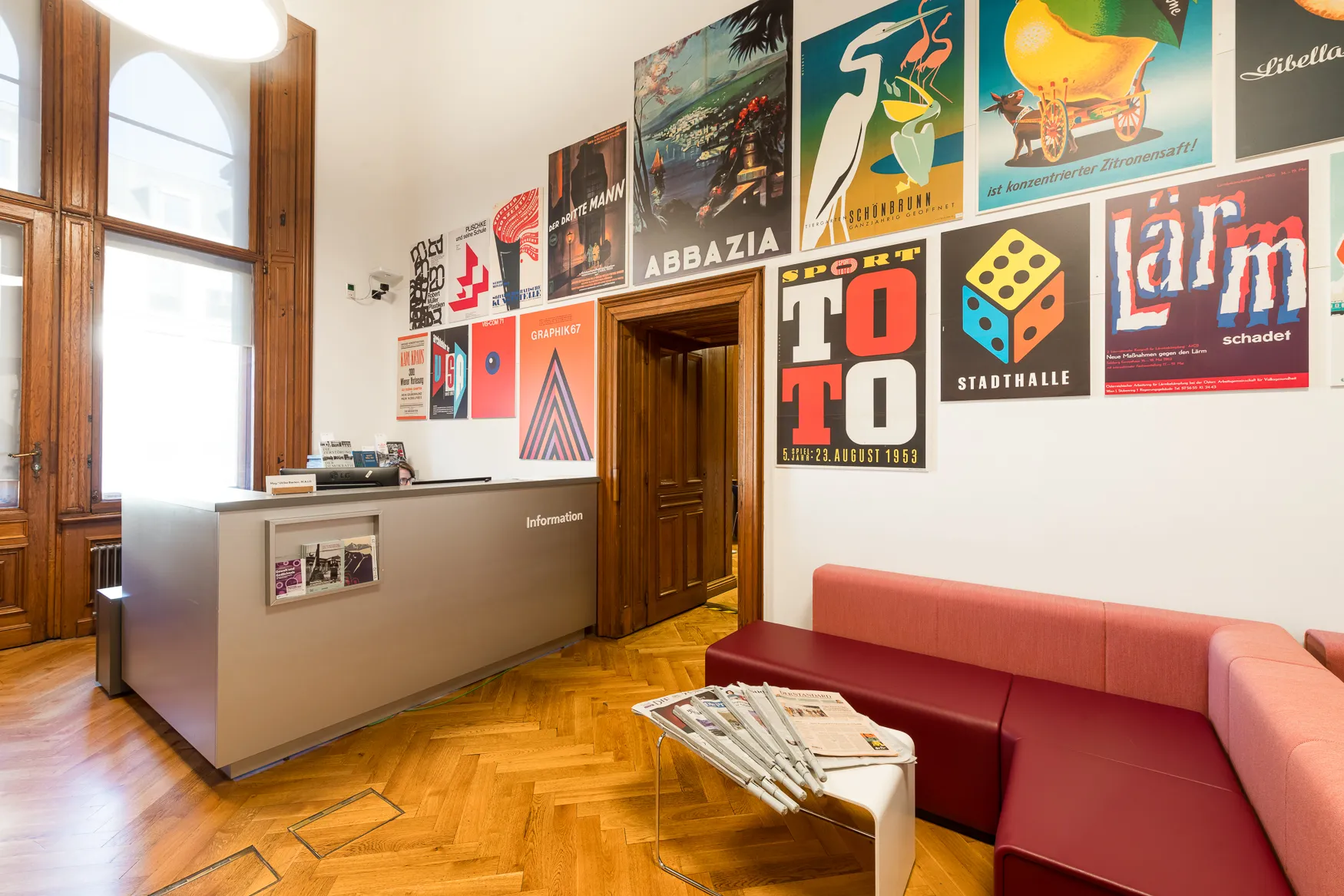 Wnętrze Wienbibliothek im Rathaus z recepcją, czerwoną kanapą i zabytkowymi plakatami na ścianie