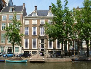 Historic brick canal house with large windows and gabled roof along a waterway, small boat docked nearby, trees lining the street