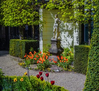 Garden with stone statue, vibrant tulips, and manicured hedges in front of a historic canal house facade