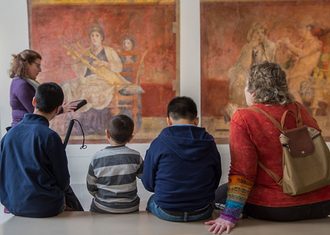 Five visitors sitting on a bench viewing two large historical paintings in a museum gallery with white walls