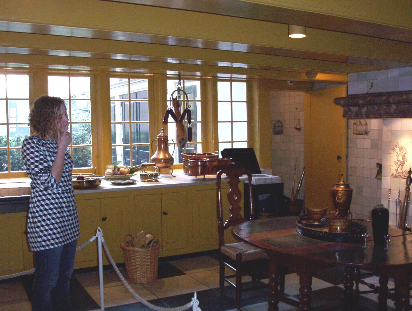 A woman in a checkered shirt stands in a historic kitchen with yellow walls, wooden furniture, and a large copper kettle on a counter.
