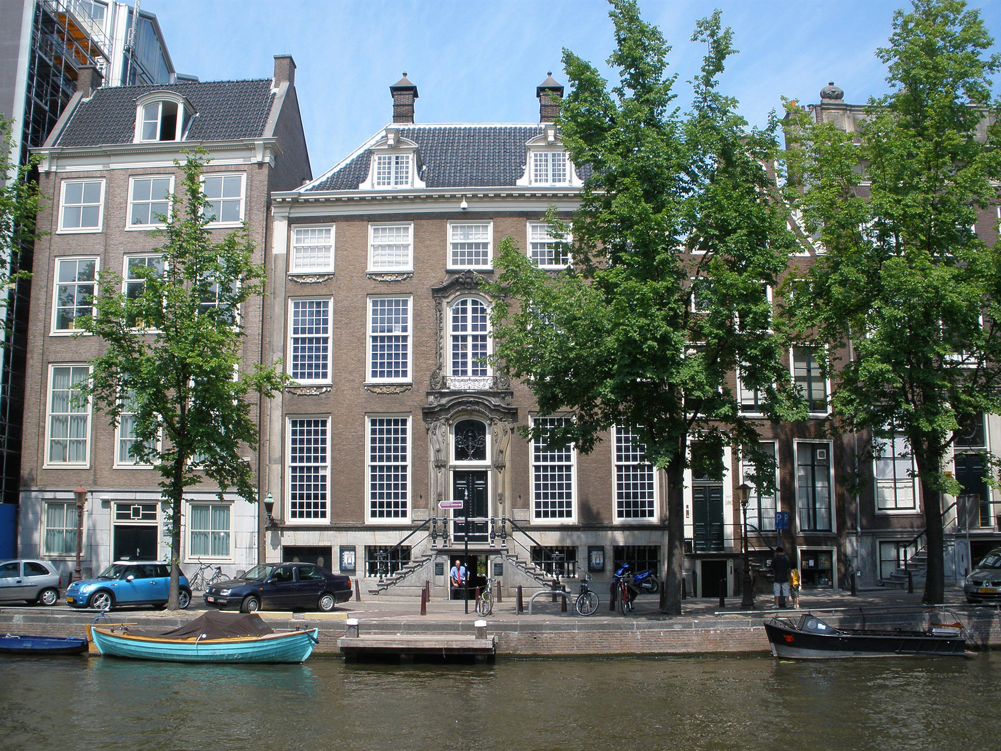 Historic canal house museum with tall windows and stone facade along a waterway, boats docked nearby and trees lining the street