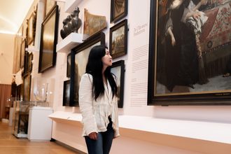 Woman standing in a museum gallery viewing framed paintings on white walls, with glass display cases visible in the background.