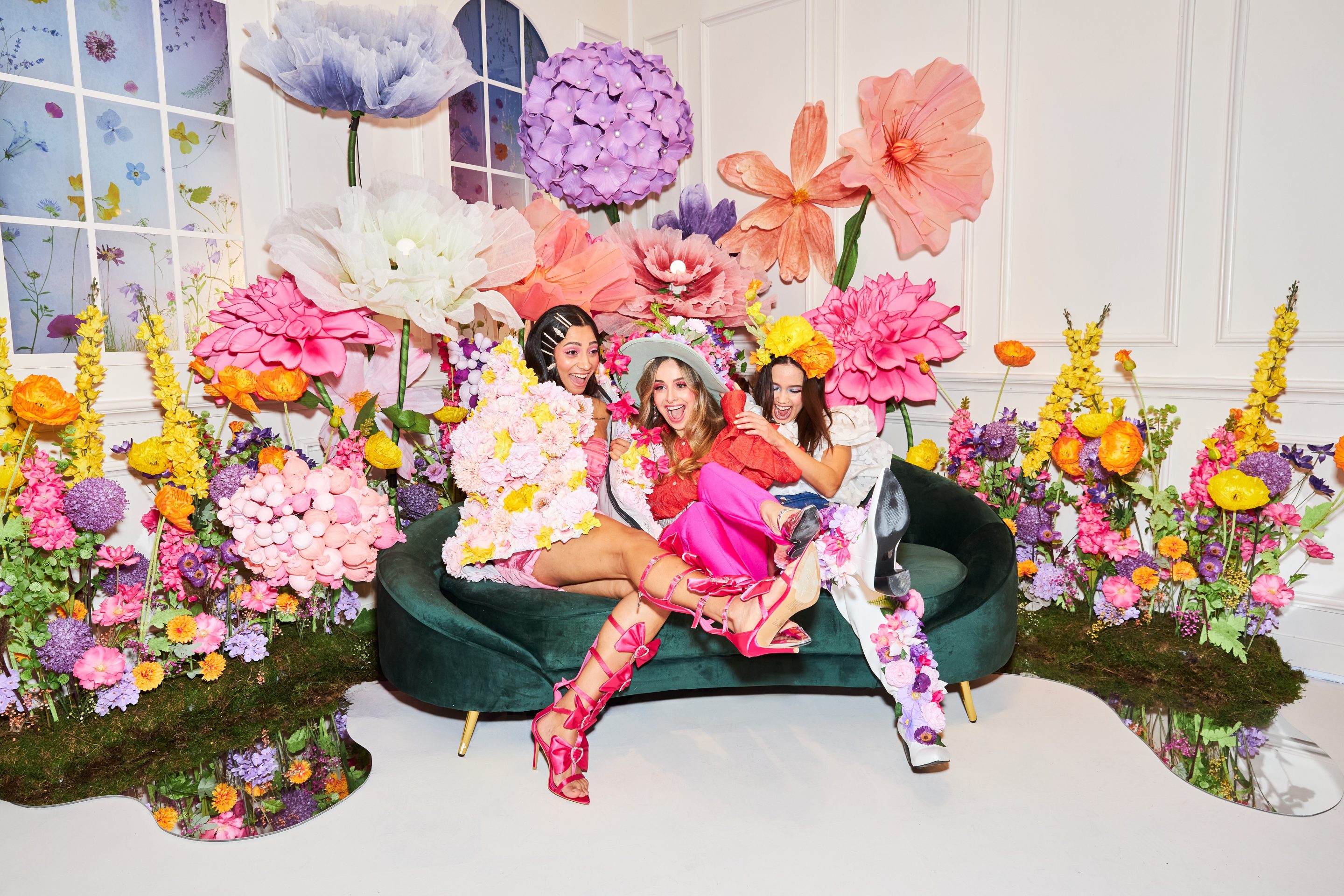 Three people seated on a green velvet couch surrounded by oversized colorful floral installations with vibrant pink, purple, and orange flowers