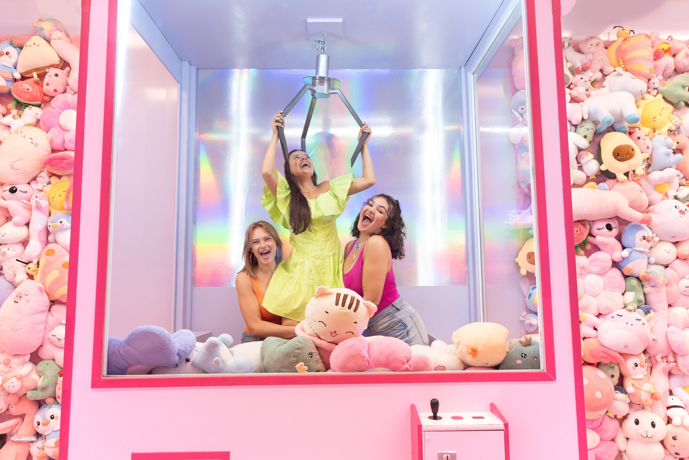Three people inside a large claw machine installation surrounded by colorful plush toys in a pink room with rainbow lighting