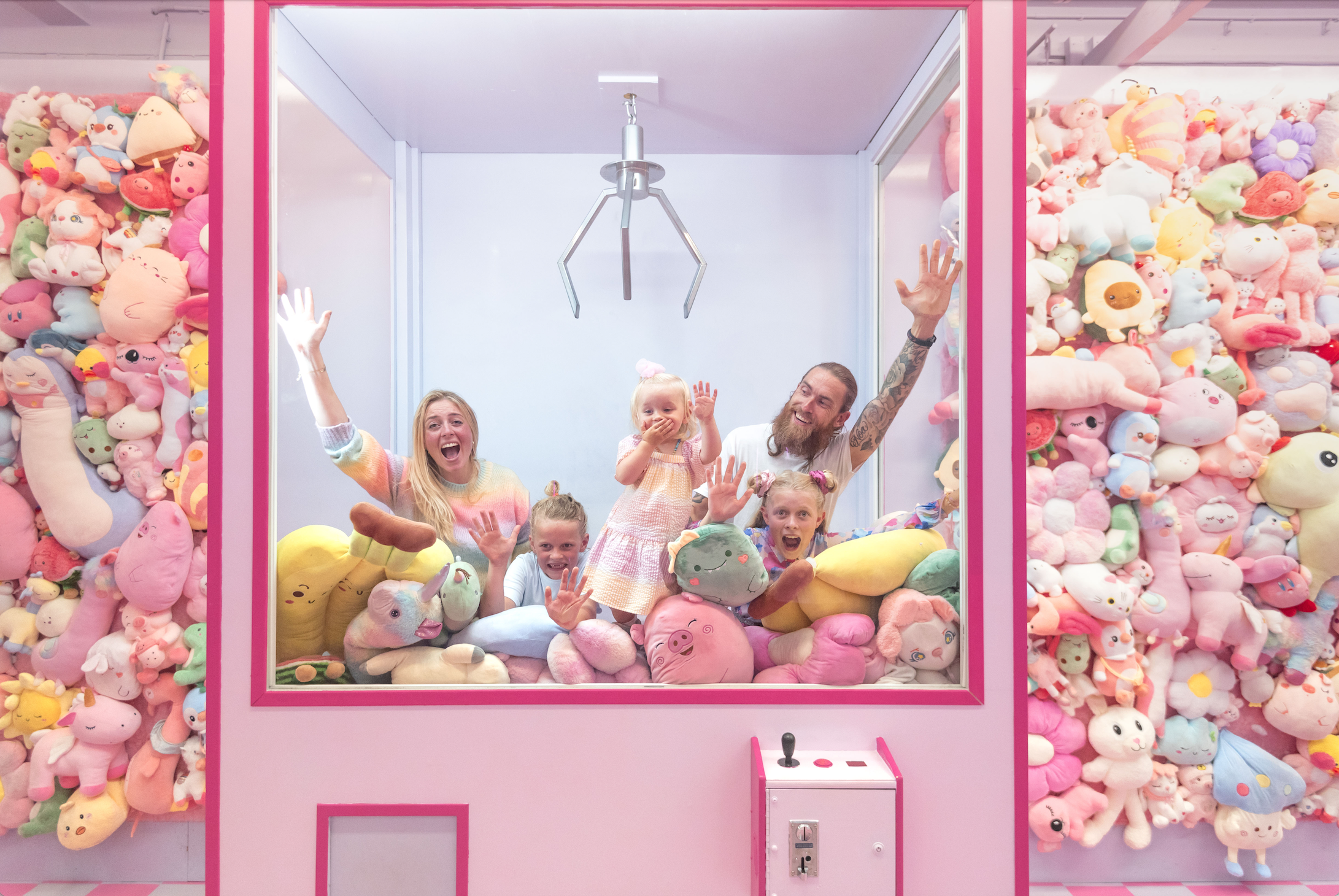Group of people inside a pink claw machine surrounded by colorful plush toys, with toy-covered walls on both sides.