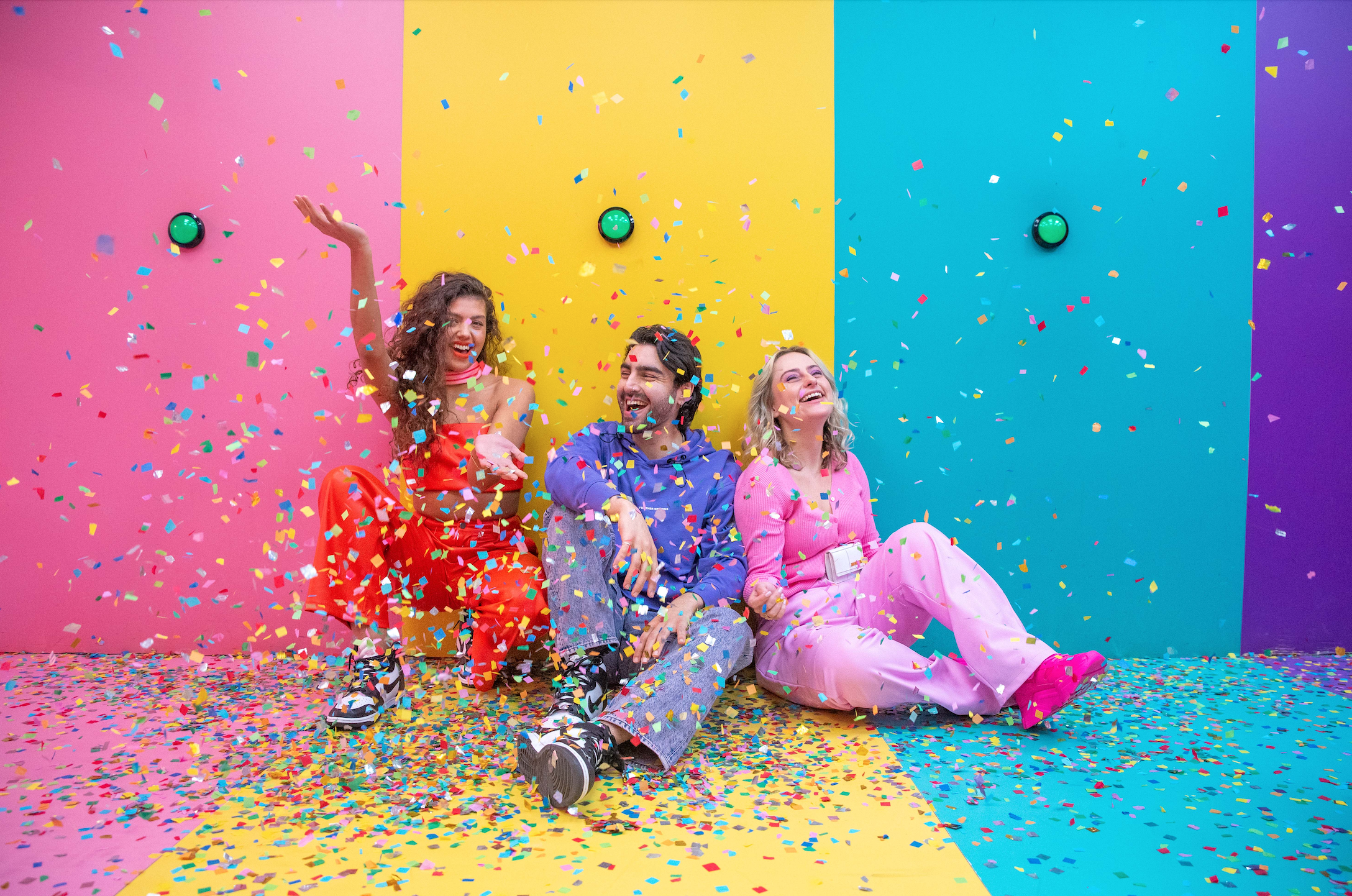 Three people sitting on a colorful floor with confetti, surrounded by pink, yellow, blue, and purple walls in an interactive exhibition space