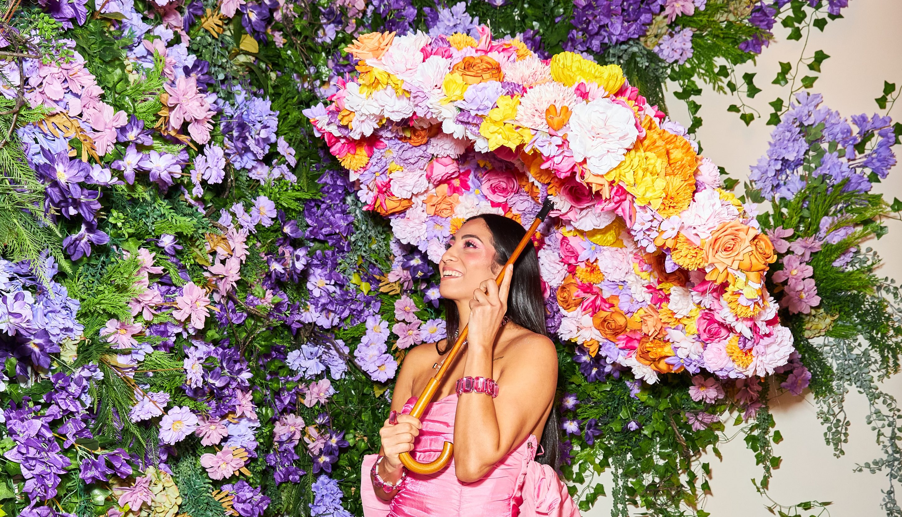 A woman in a pink dress holding a large, colorful floral umbrella stands in front of a vibrant wall covered in purple and pink flowers.