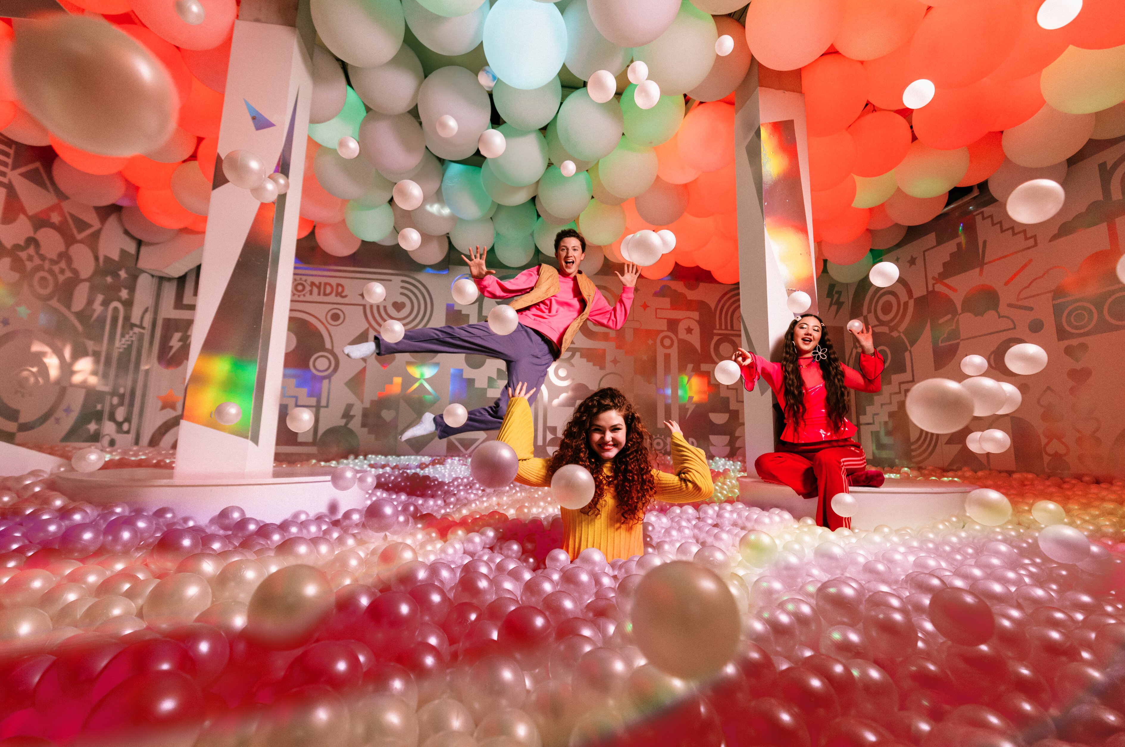 Three people posing joyfully among colorful balloons and vibrant patterned walls in an interactive art installation space