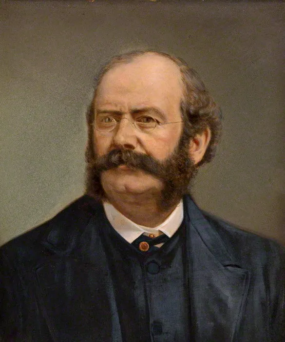Portret łysiejącego mężczyzny z gęstym, ciemnym wąsem i brodą, ubranego w okulary w cienkich oprawkach i ciemnoniebieski garnitur.