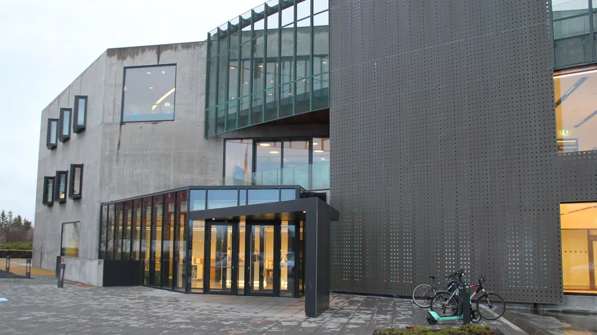 Nowoczesny budynek muzeum z elewacją z ciemnego, perforowanego metalu, szklanym zadaszeniem wejścia z ciepłym oświetleniem wnętrza oraz rowerami zaparkowanymi na wybrukowanym placu przed budynkiem.