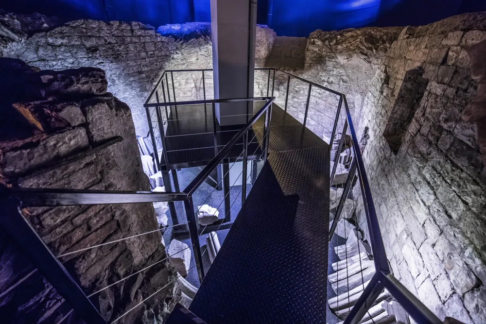 Modern metal staircase with grated platform inside ancient stone walls under blue lighting