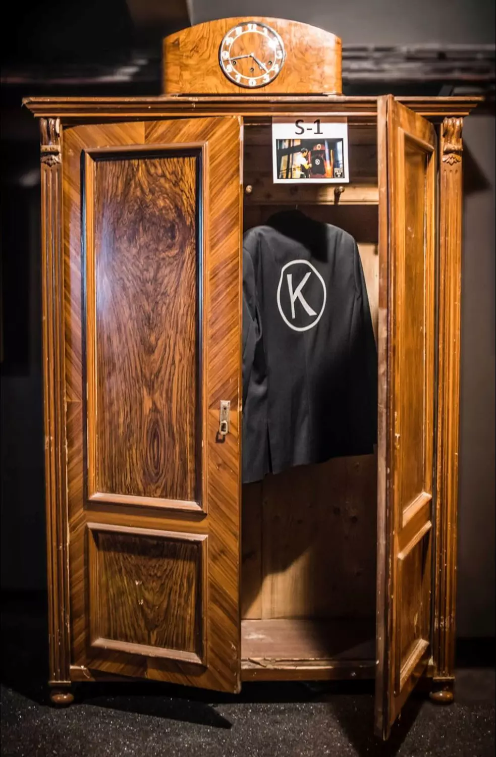 An open wooden wardrobe with a clock on top and a black jacket with a 'K' logo hanging inside