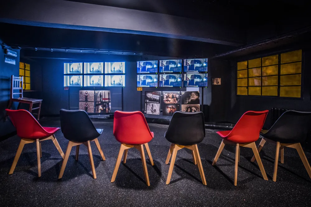 A dark exhibition room with red and black chairs arranged in a circle facing multiple screens displaying multimedia content.