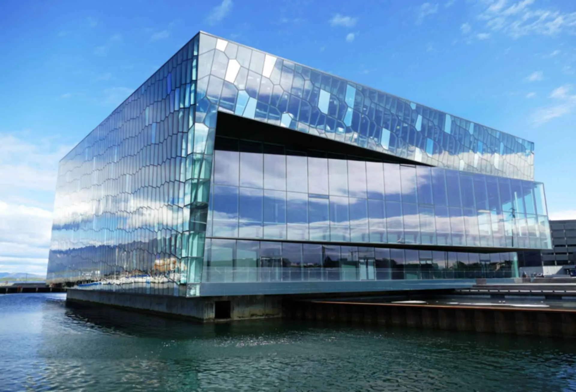 Un gran edificio moderno de cristal con una fachada geométrica que refleja el cielo azul y las nubes, situado a orillas de un puerto.