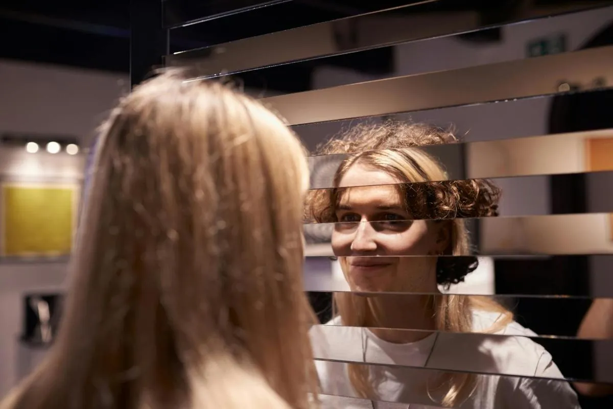Una persona con el pelo rubio mirando un espejo con lamas horizontales que distorsionan su reflejo