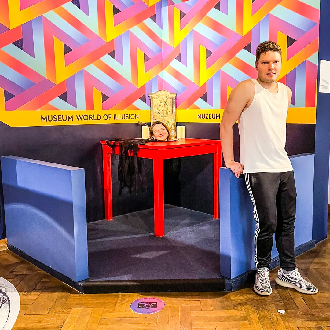 Man standing next to an optical illusion exhibit with a woman appearing to float above a red table, in front of a colorful geometric wall with 'Museum World of Illusion' text