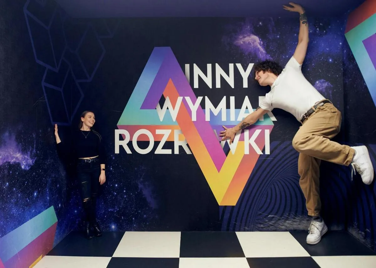 Two people interacting with an optical illusion exhibit featuring a checkered floor and a colorful backdrop with text