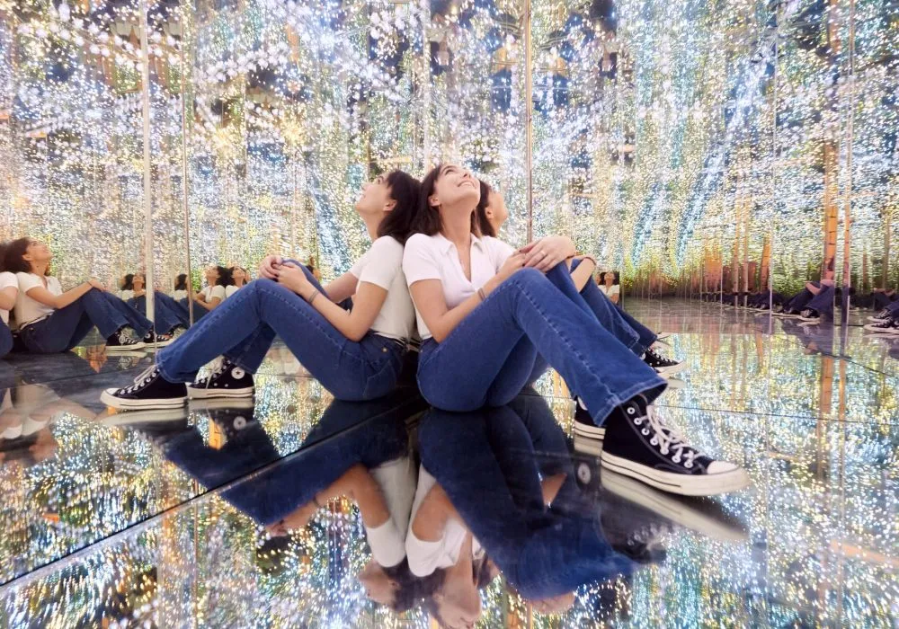 Two people sitting back-to-back on a reflective floor surrounded by mirrored walls with light patterns in a mirrored room