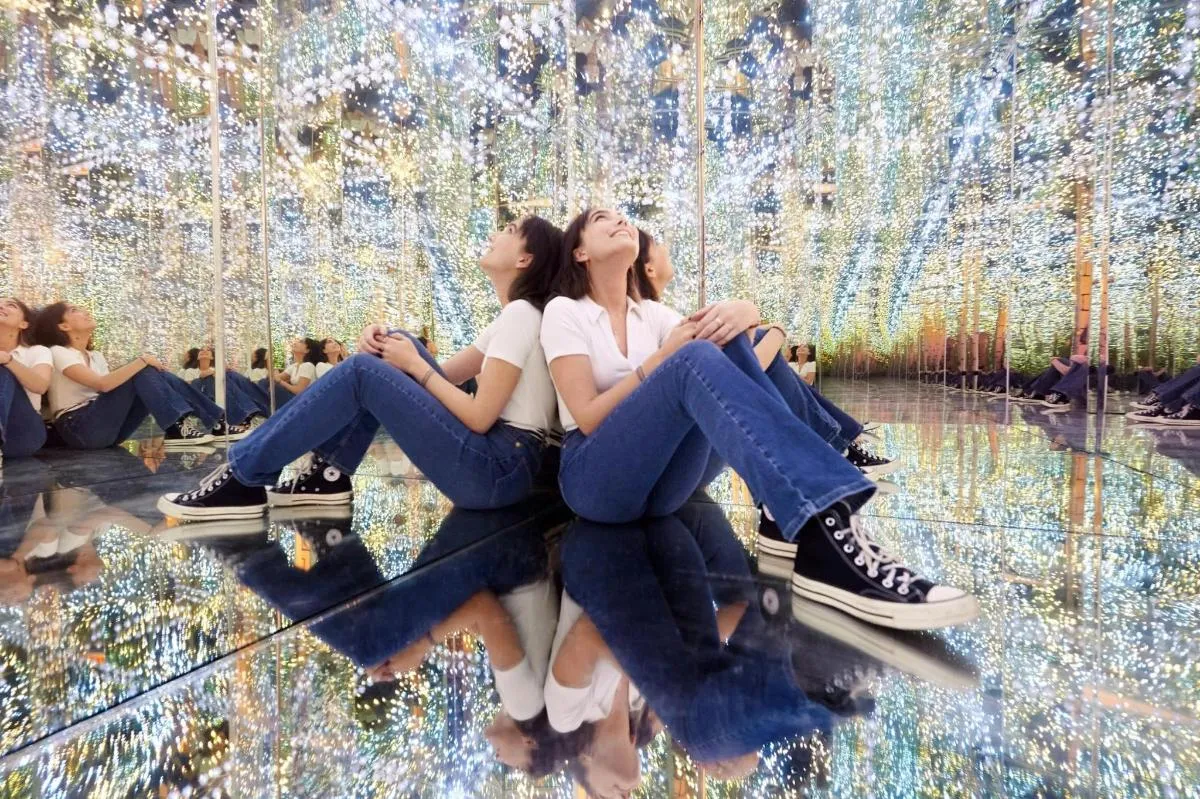 People sitting on a reflective floor surrounded by a ceiling of sparkling lights in an optical illusion exhibit