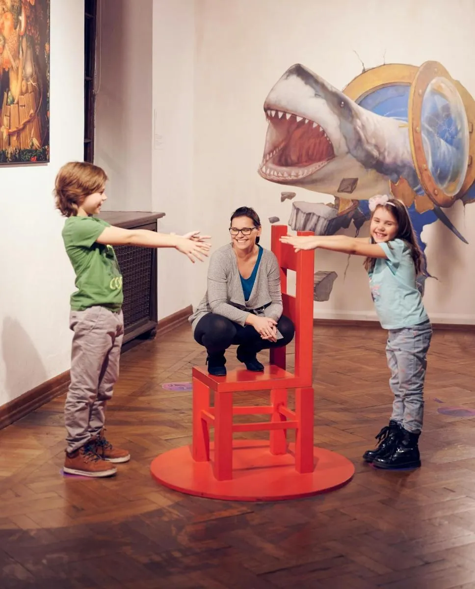 Niños y un adulto interactuando con una ilusión óptica de una silla roja frente a un mural de tiburón