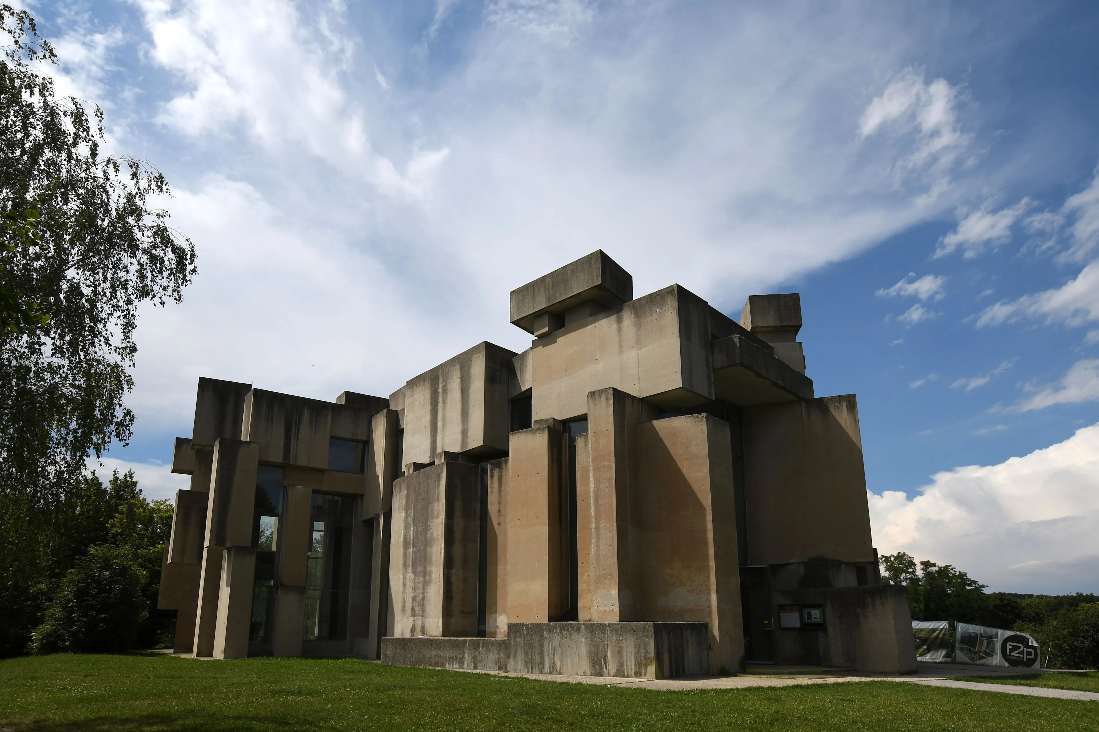 Moderne betonnen kerkgebouw met geometrische gestapelde blokken, omgeven door groen gras en bomen onder een blauwe lucht met wolken
