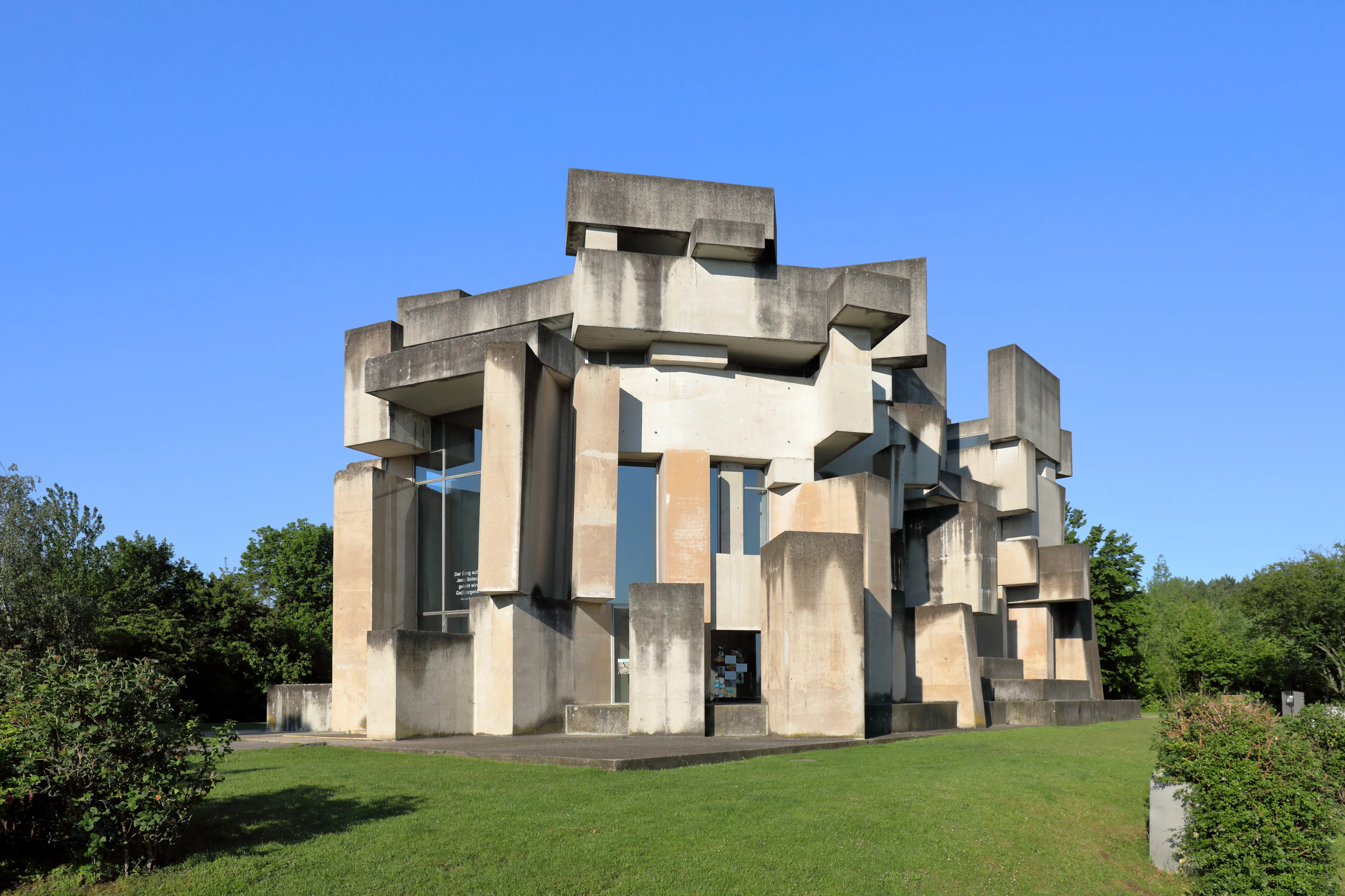 Een moderne betonnen kerk met geometrisch gestapelde blokken en grote ramen, tegen een blauwe lucht en groen gras.