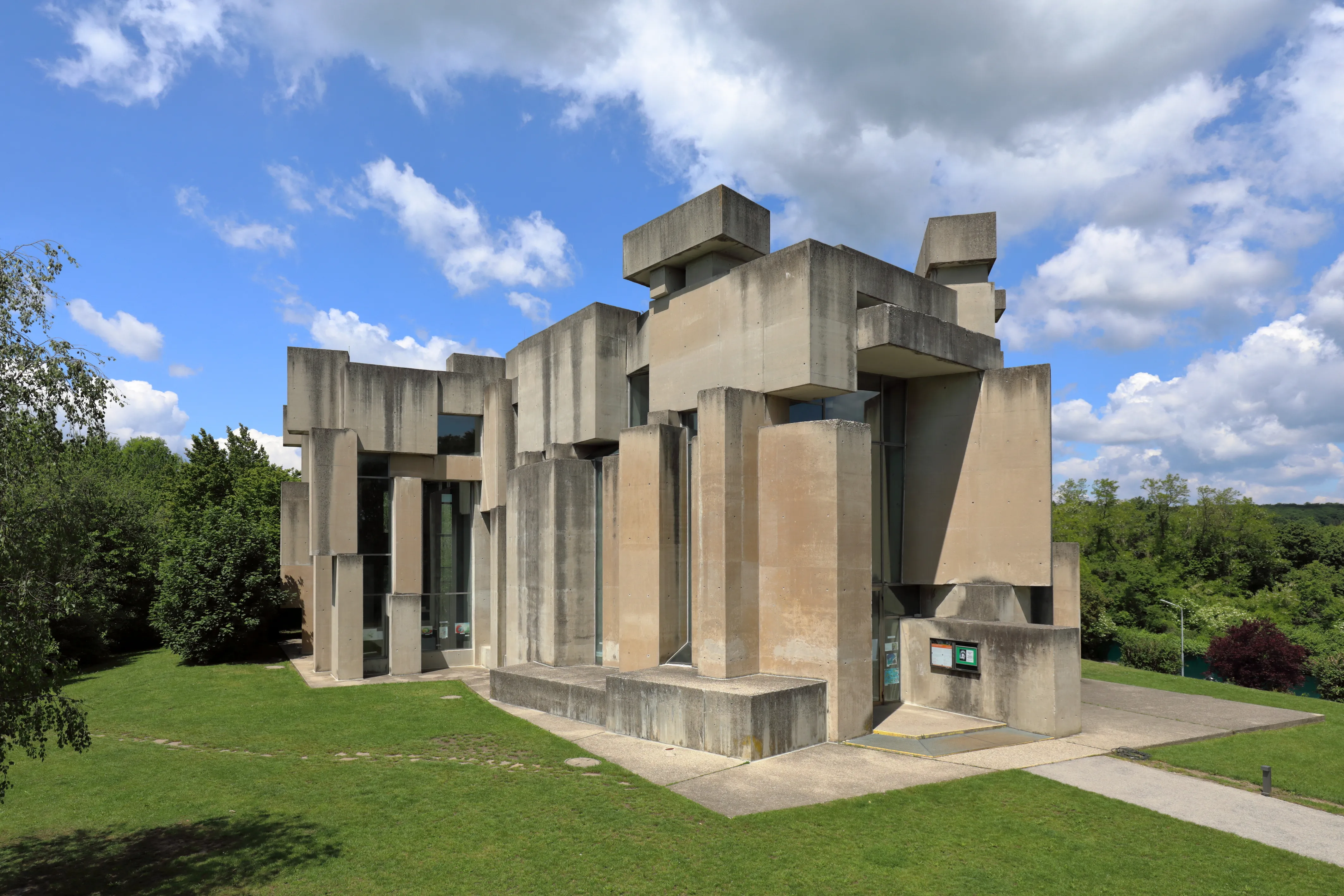 Moderne betonnen kerk met geometrische gestapelde blokken, omringd door groen gras en bomen onder een blauwe lucht met wolken