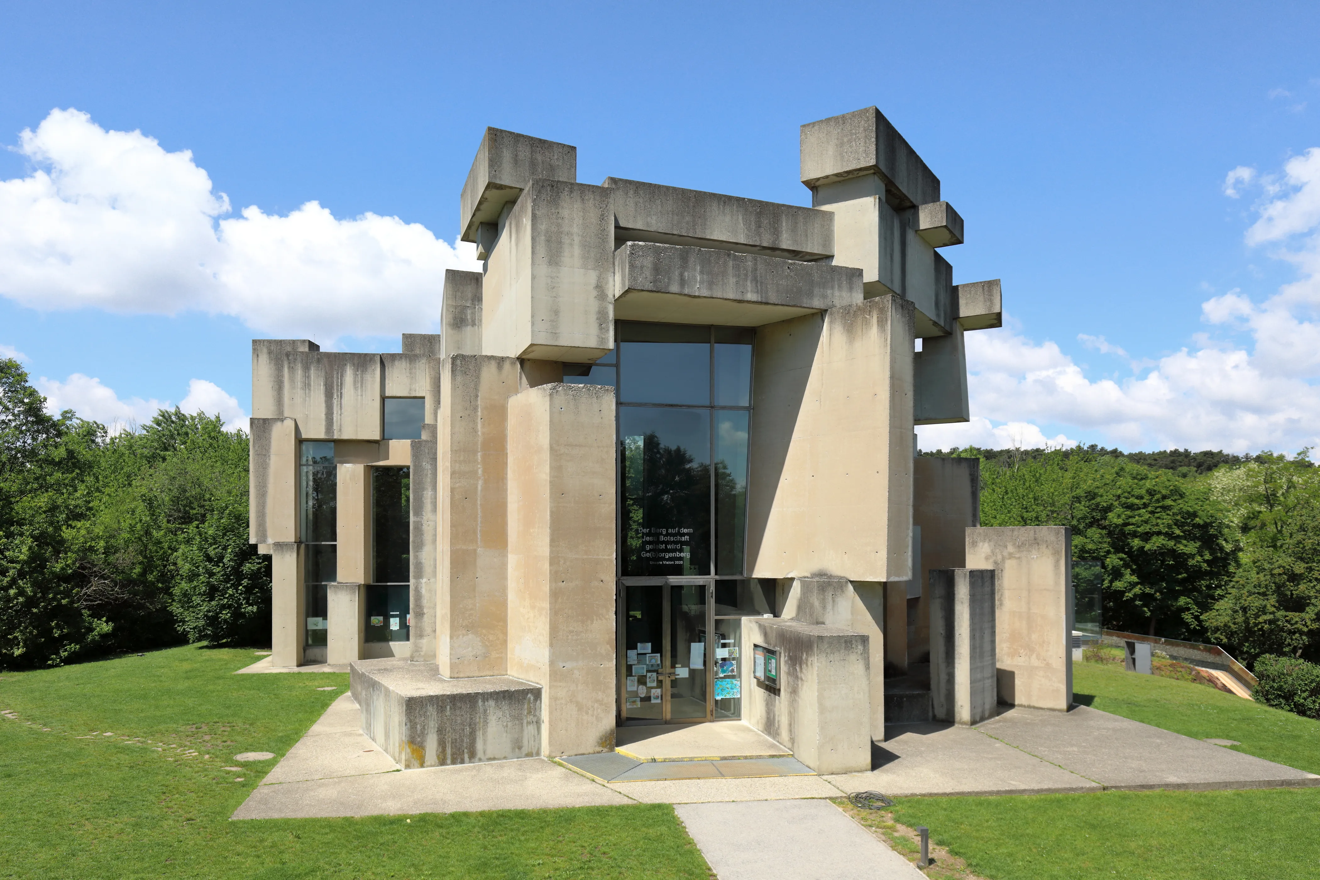 Modern betonnen kerkgebouw met geometrische blokken en grote glazen ingang op een grasveld