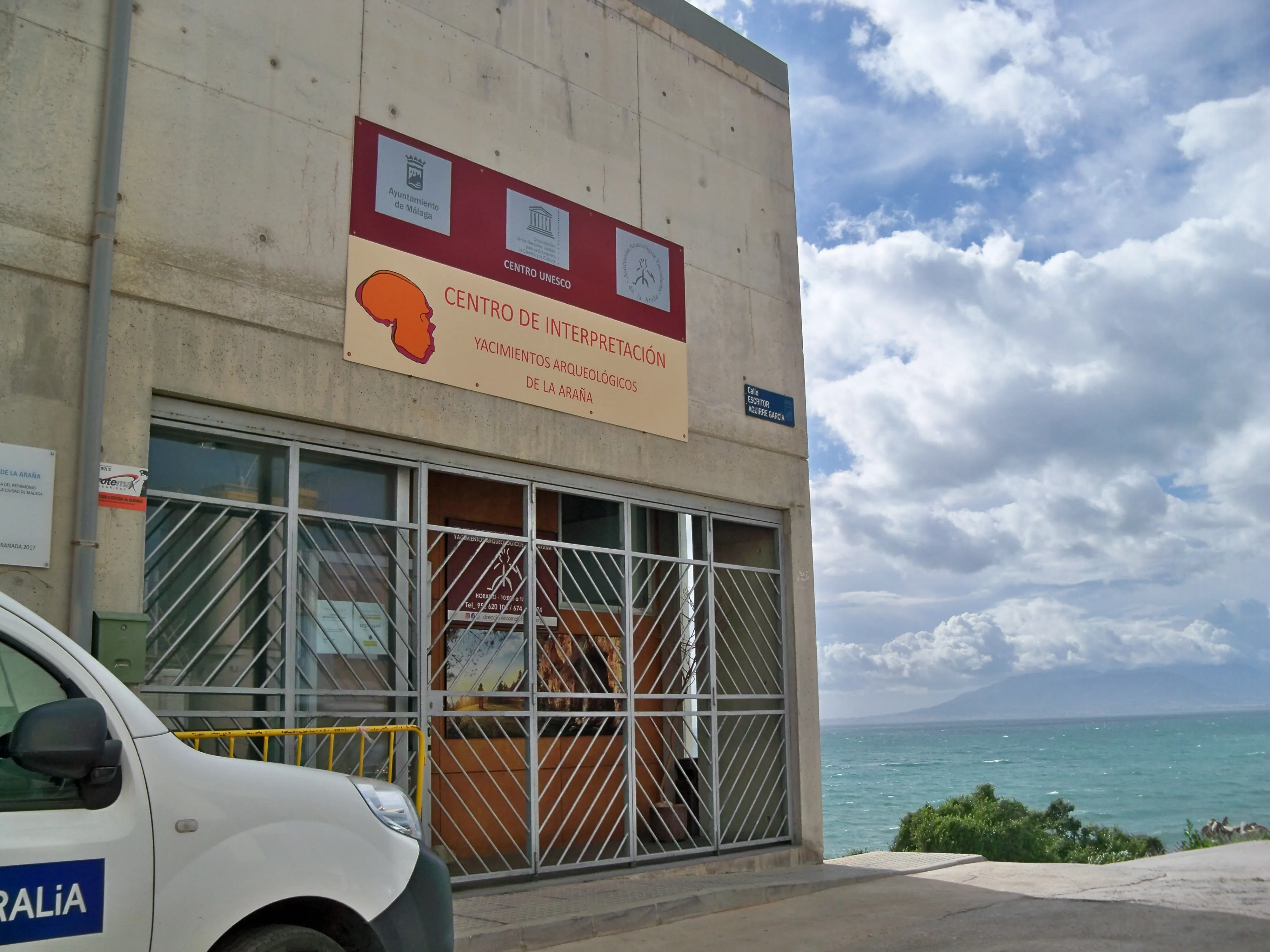 Exterior de un edificio de hormigón con un letrero grande que dice 'Centro de Interpretación de los Yacimientos Arqueológicos de La Araña' y un cartel rojo encima, con rejas de seguridad metálicas en las puertas de cristal y el mar visible al fondo bajo un cielo nublado.