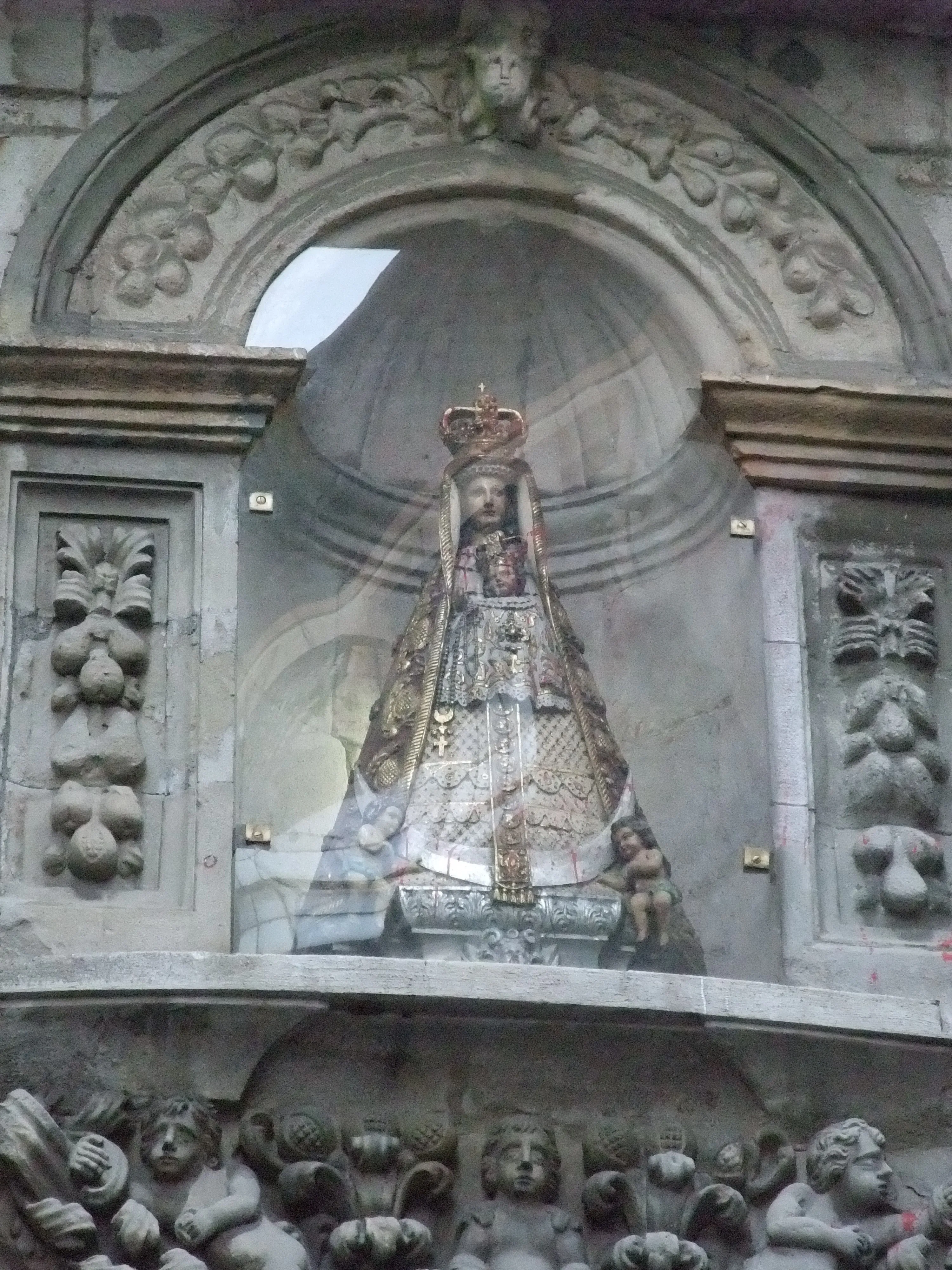 Stone niche on a Baroque facade containing a crowned statue of a religious figure behind glass, flanked by carved panels and cherub figures below