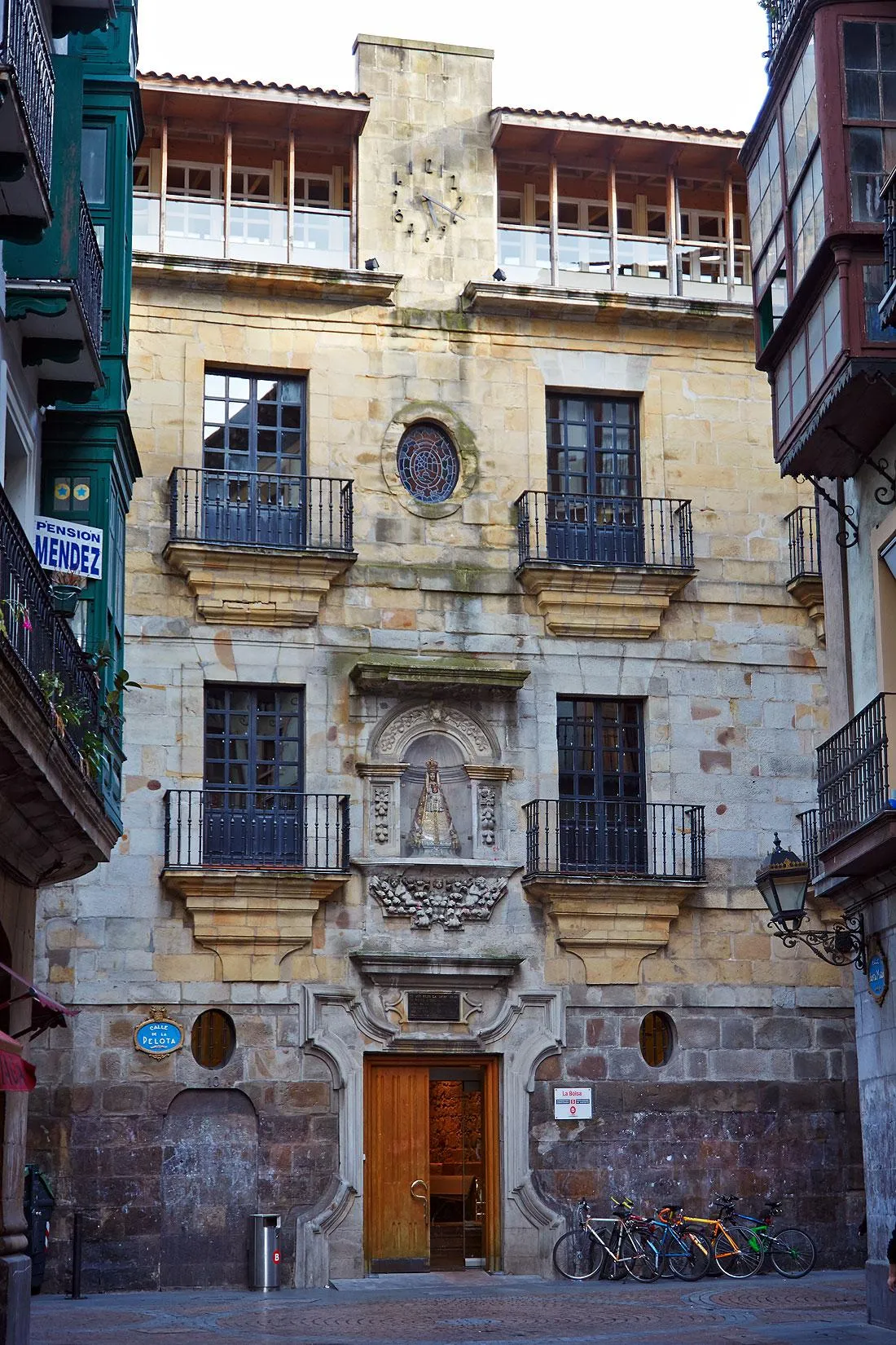 Stone facade of the Yohn Palace La Bolsa featuring iron balconies, a central statue niche, and a wooden entrance door with bicycles parked outside.