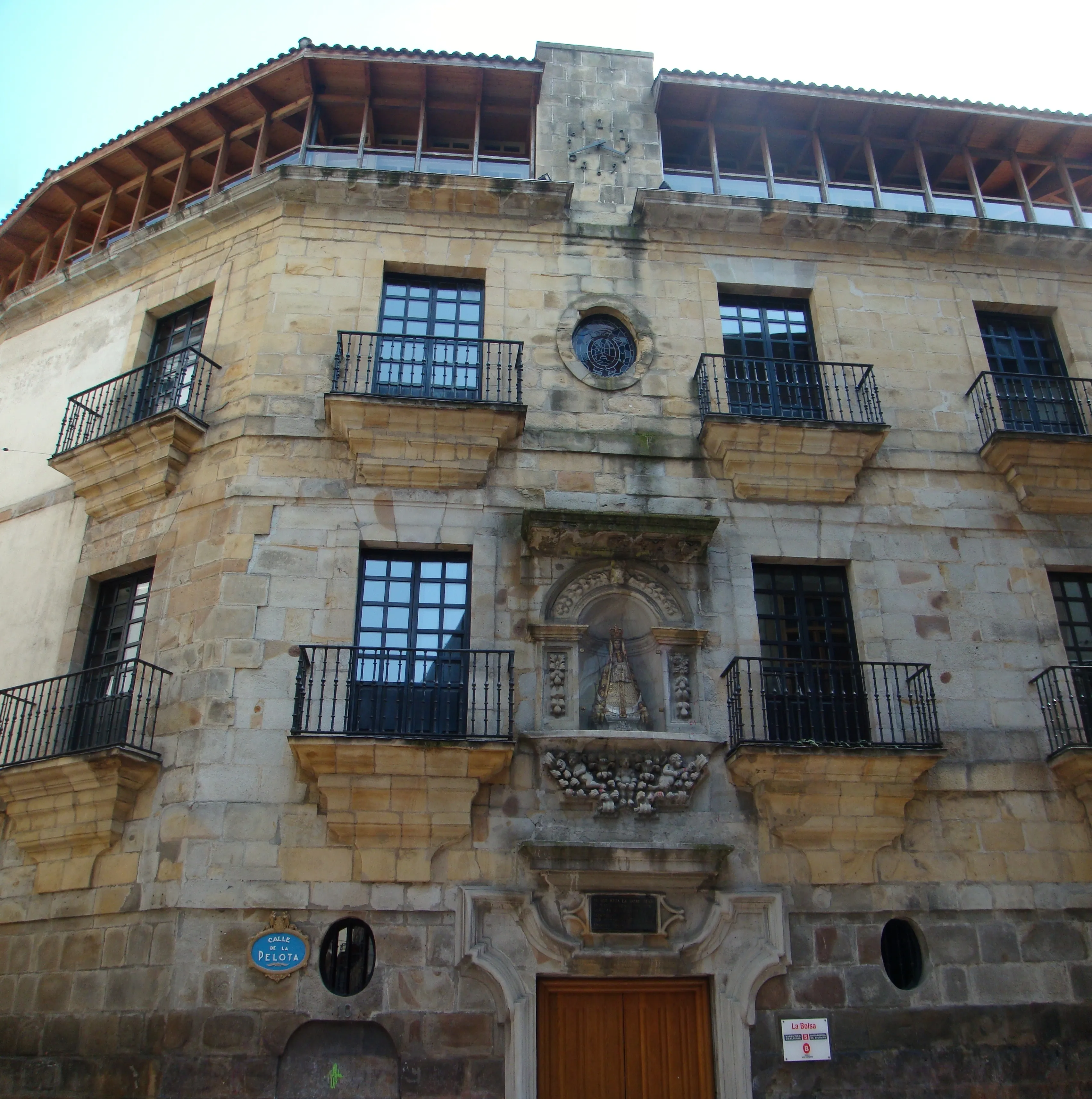 Stone Baroque palace facade with iron balconies, arched doorway with decorative niche, and wooden roof overhang at the corner of the building