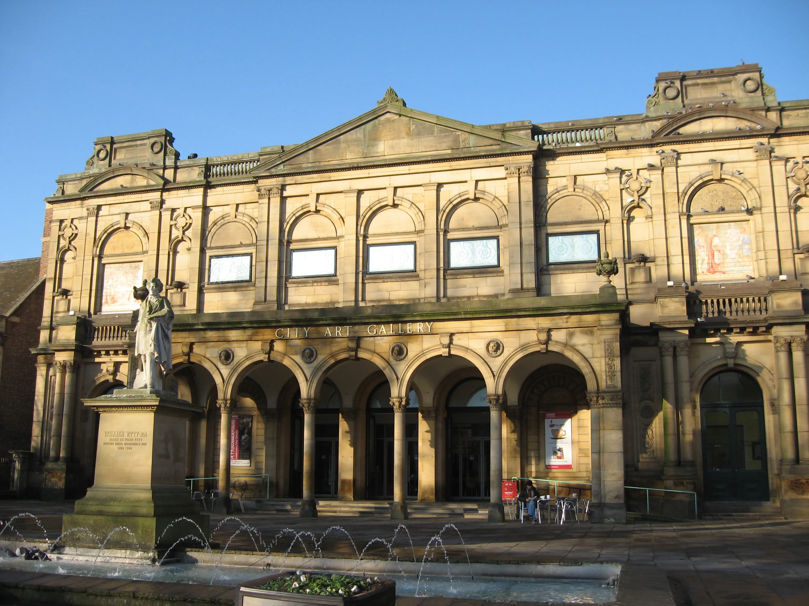 Kamienna fasada York Art Gallery z łukowymi oknami, kolumnadą na parterze, posągiem na cokole i fontanną przed wejściem na tle bezchmurnego, błękitnego nieba
