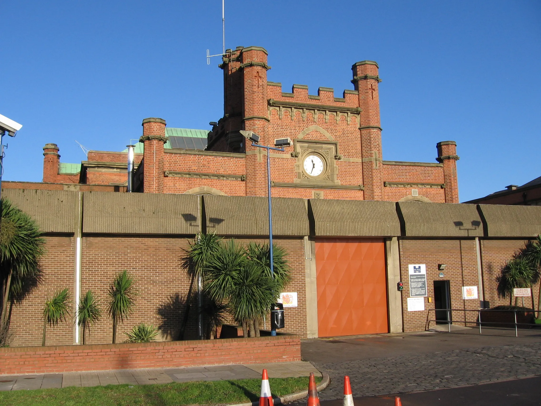 Exterior de un edificio penitenciario de ladrillo rojo con una torre del reloj, almenas y una gran puerta naranja bajo un cielo azul despejado