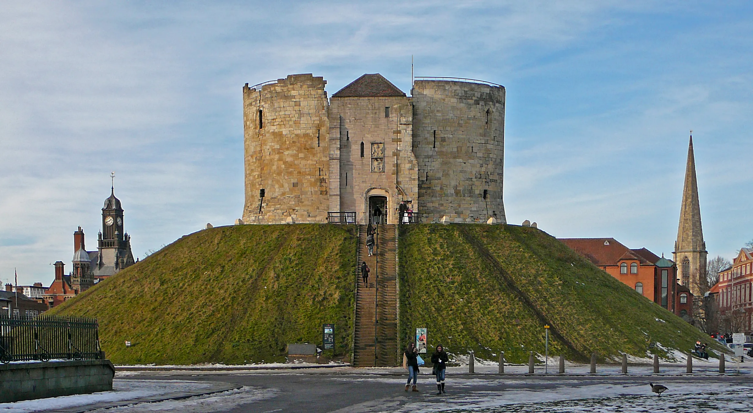 Clifford's Tower, una torre medieval de piedra sobre un montículo de hierba, con personas caminando a sus pies y torres de iglesias visibles al fondo bajo un cielo azul