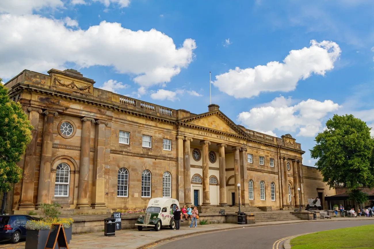 Vista exterior del York Castle Museum, un gran edificio neoclásico de piedra arenisca con columnas y un frontón, bajo un cielo azul con nubes blancas, con una furgoneta blanca clásica aparcada delante.
