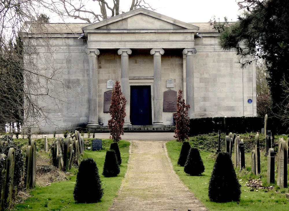 Neoklassieke stenen kapel met zuilen en fronton op York Cemetery, gezien vanaf een pad omzoomd door conische groenblijvende struiken en grafstenen