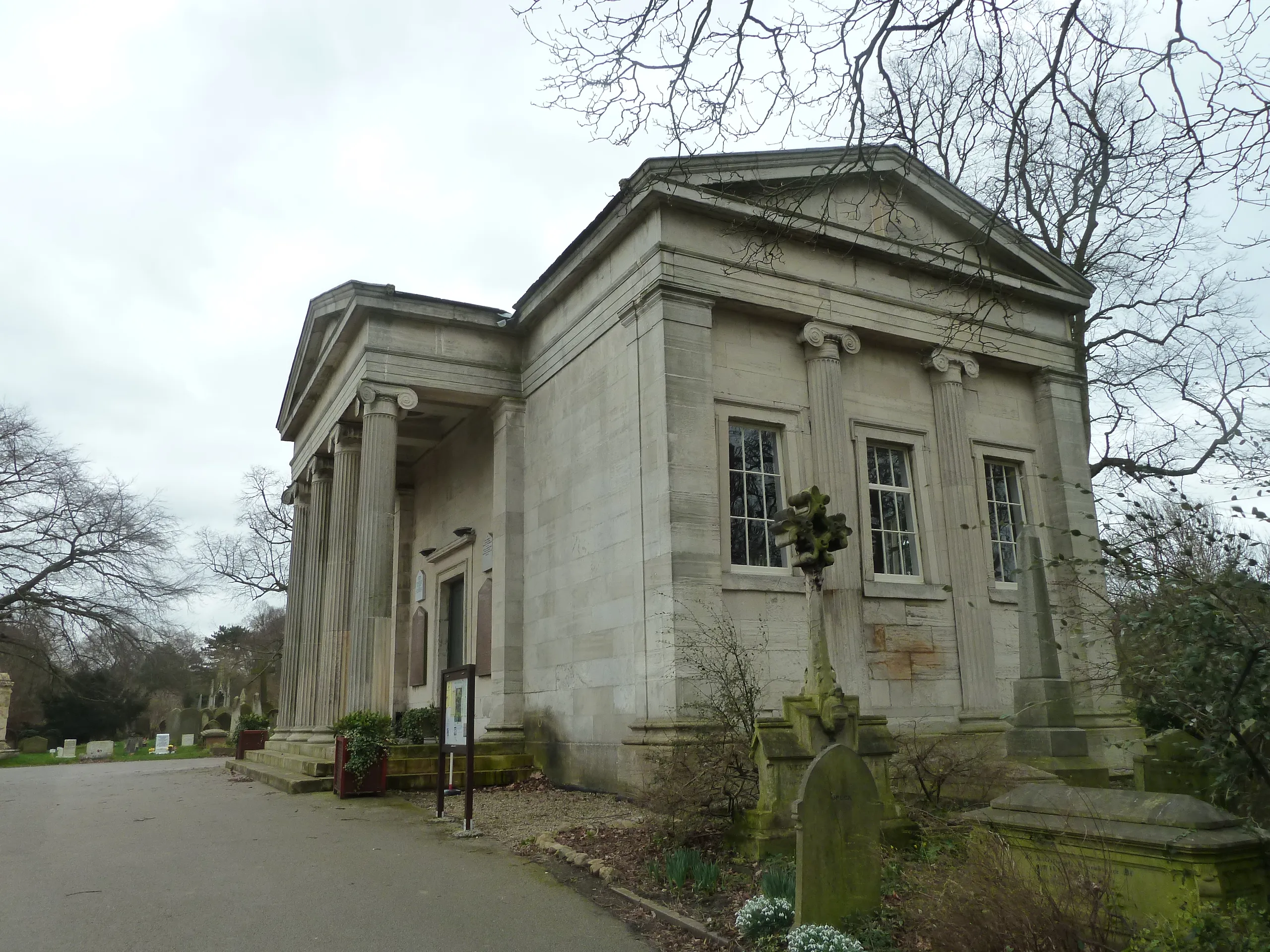 Klassiek stenen gebouw met zuilen en een fronton op York Cemetery, met grafstenen op de voorgrond en kale bomen.