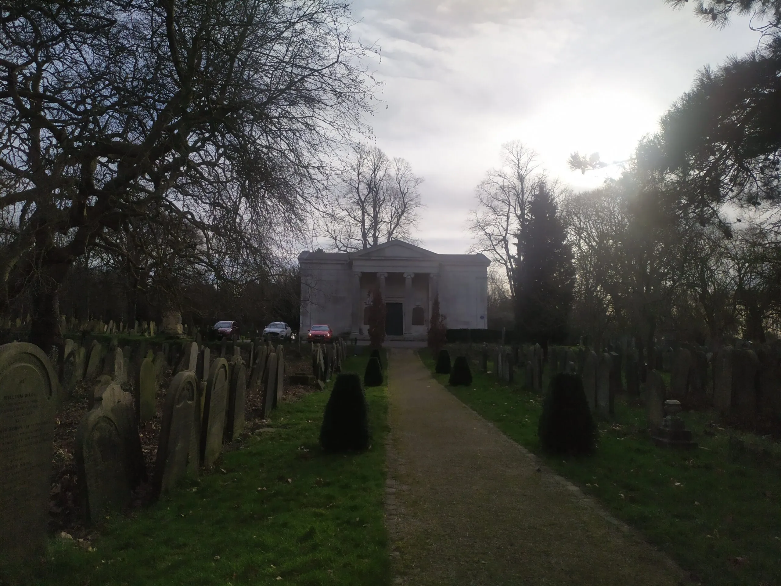 Een pad omzoomd met grafstenen leidt naar een klassiek wit gebouw op York Cemetery, waarbij kale bomen afsteken tegen een heldere, bewolkte lucht.