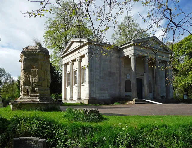 Neoklassiek stenen gebouw met zuilen en fronton in een groene begraafplaatsomgeving, met links een verweerd stenen monument.