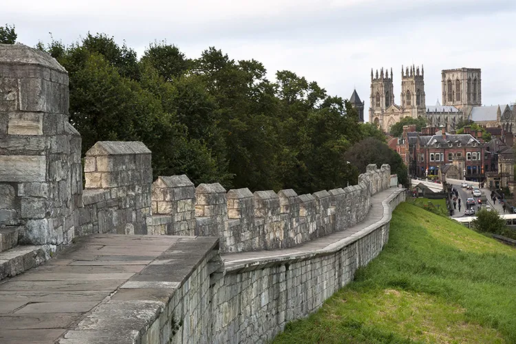 Camino de piedra a lo largo de las York City Walls con almenas; las torres de York Minster son visibles al fondo entre los árboles.