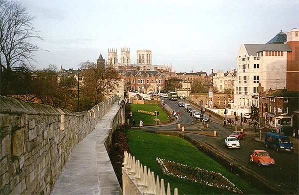 El camino de piedra a lo largo de las York City Walls se extiende en la distancia con las torres de York Minster visibles en el horizonte, dominando una carretera con coches y céspedes verdes abajo.