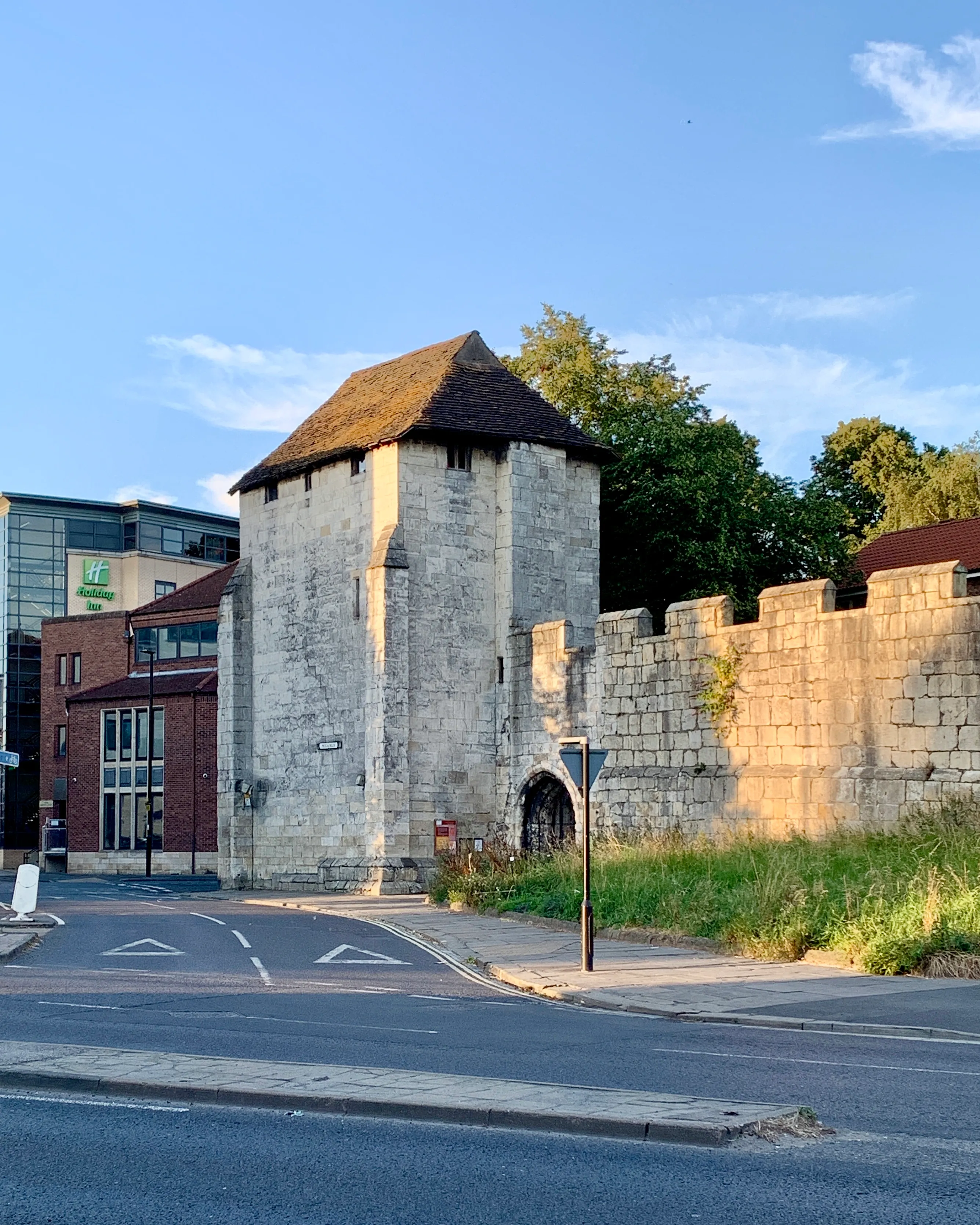 Torre de piedra medieval con techo de tejas oscuras conectada a un muro de piedra almenado, con un hotel moderno visible a la izquierda y una carretera en primer plano.