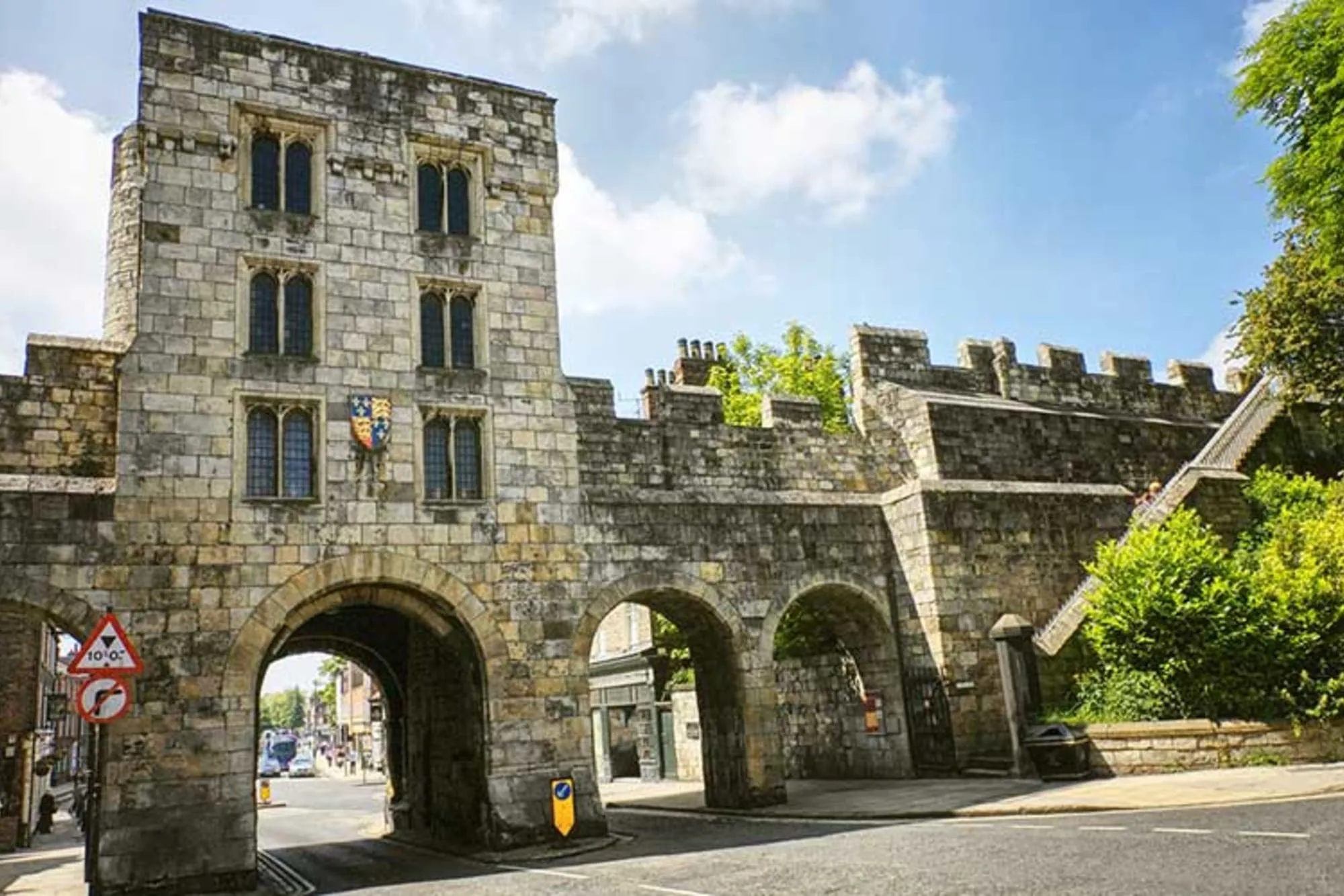 Casa de guardia de piedra medieval con múltiples arcos y almenas que forma parte de las murallas de York, con una carretera que pasa a través del arco principal.