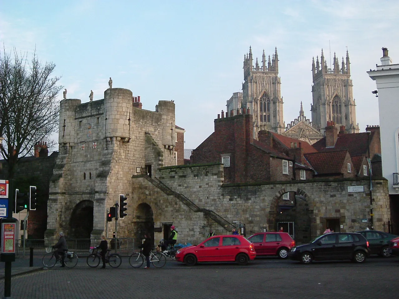 Casa de guardia medieval de piedra con entrada arqueada, ciclistas y coches en la calle, con las torres gemelas de York Minster al fondo.